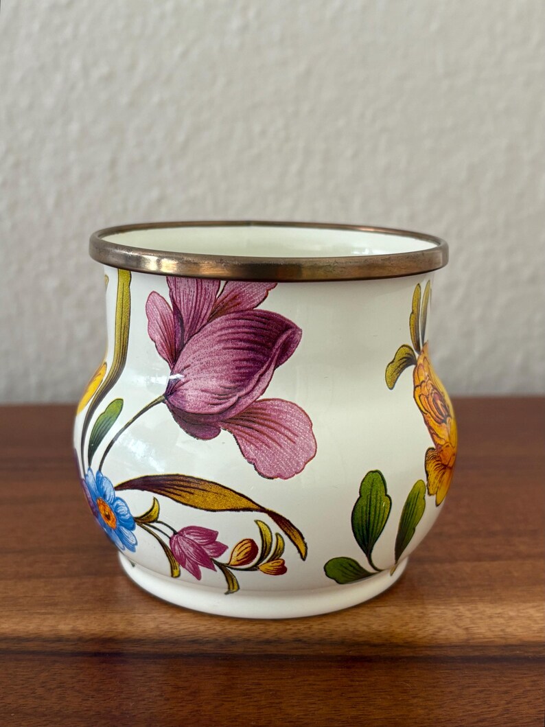 May include: A small, white ceramic vase with a floral design. The vase features a brass rim and is adorned with purple, blue, and yellow flowers with green leaves. The flowers are painted on the exterior of the vase.