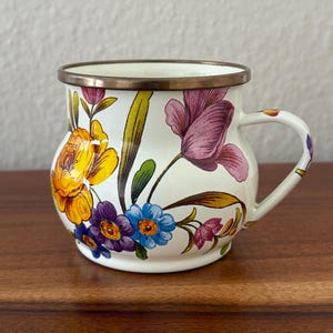 May include: A white enamel mug with a floral design featuring yellow, purple, and blue flowers. The mug has a bronze-colored rim and handle. The flowers are painted in a detailed, colorful style, creating a vintage aesthetic.