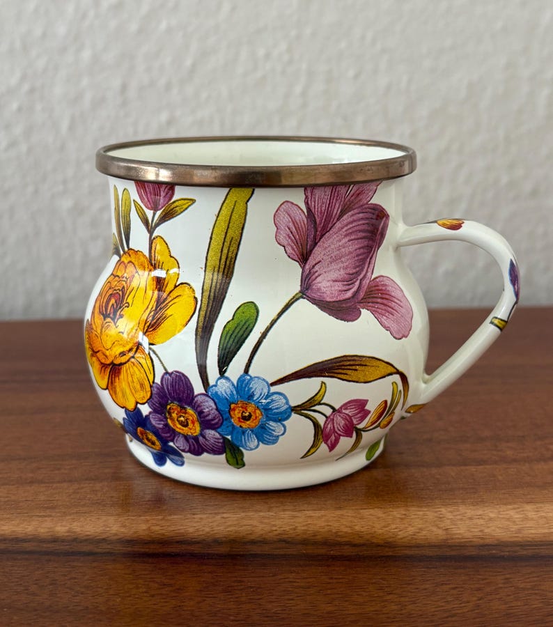 May include: A white enamel mug with a floral design featuring yellow, purple, and blue flowers. The mug has a bronze-colored rim and handle. The flowers are painted in a detailed, colorful style, creating a vintage aesthetic.