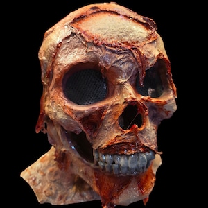 May include: A full-head skull mask featuring a textured, fleshy appearance with shades of tan, brown, and deep red. The mask includes hollow eye sockets covered with black mesh, a nasal cavity, and a set of exposed, realistic-looking teeth.
