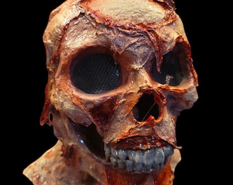 May include: A full-head skull mask featuring a textured, fleshy appearance with shades of tan, brown, and deep red. The mask includes hollow eye sockets covered with black mesh, a nasal cavity, and a set of exposed, realistic-looking teeth.