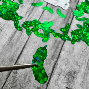 May include: A green glitter pickle shaped embellishment. The pickle is made of iridescent green glitter and is about 2 inches long. The pickle is being held by tweezers.