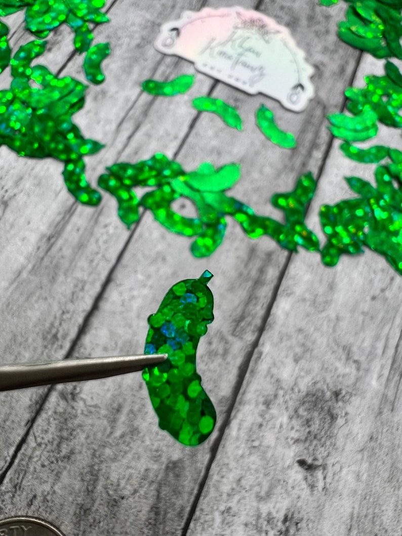 May include: A green glitter pickle shaped embellishment. The pickle is made of iridescent green glitter and is about 2 inches long. The pickle is being held by tweezers.