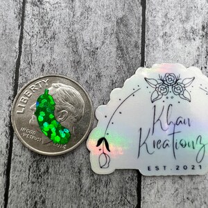 May include: A green and silver glitter sticker with a green pickle design. The sticker is on a white background with a holographic finish. The sticker also has black text that reads "Khan Kreations EST. 2021".
