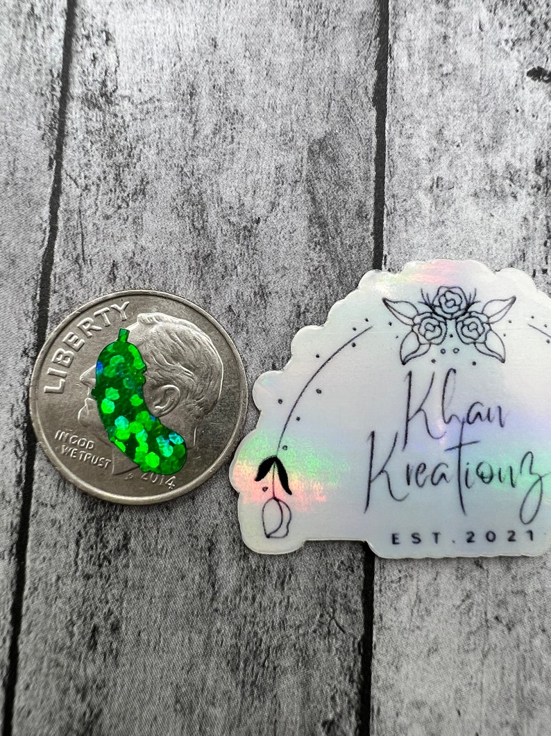 May include: A green and silver glitter sticker with a green pickle design. The sticker is on a white background with a holographic finish. The sticker also has black text that reads "Khan Kreations EST. 2021".