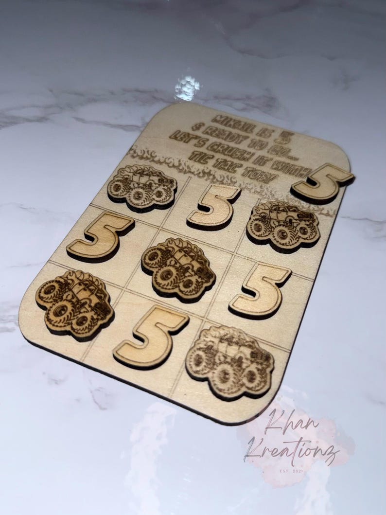 May include: Wooden tic-tac-toe game with laser-cut monster truck and number 5 designs. The game board has the text "MIKAEL IS 5 & READY TO GO... LET'S CRUSH IT WITH TIC TAC TON". Includes 9 game pieces.