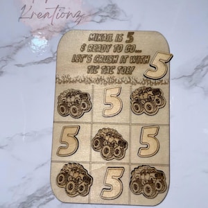 May include: Wooden Tic-Tac-Toe game with the text "MIKAIL IS 5 & READY TO GO... LET'S CRUSH IT WITH TIC TAC TOE!". The game board features monster truck and number 5 designs. A separate number 5 is included.