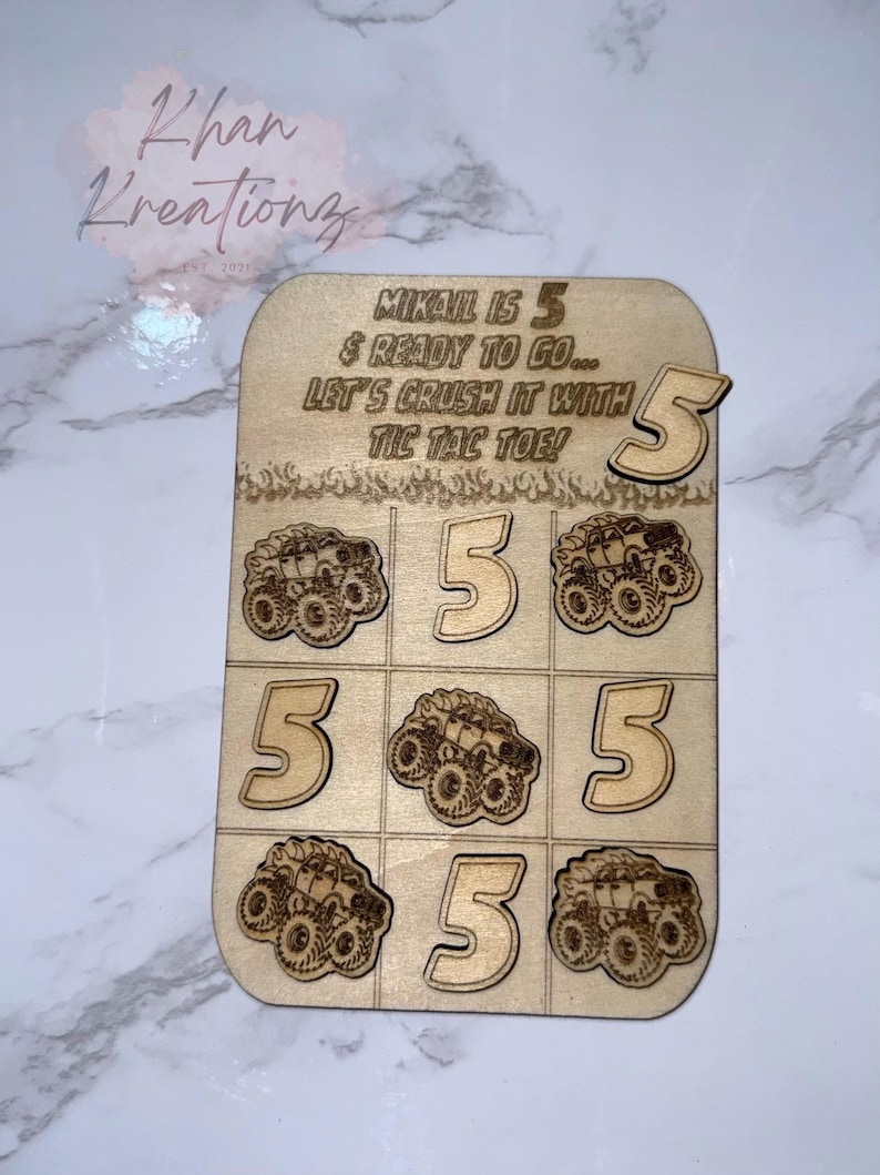 May include: Wooden Tic-Tac-Toe game with the text "MIKAIL IS 5 & READY TO GO... LET'S CRUSH IT WITH TIC TAC TOE!". The game board features monster truck and number 5 designs. A separate number 5 is included.