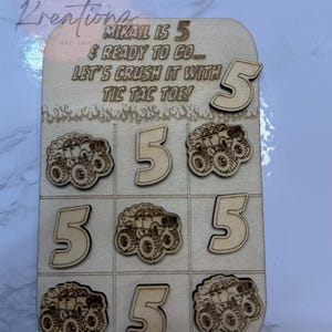 May include: Wooden tic-tac-toe game with laser-cut monster truck and number 5 designs. The game board includes the text "MIKAIL IS 5 & READY TO GO... LET'S CRUSH IT WITH TIC TAC TOE!" and the Khan Kreations logo.