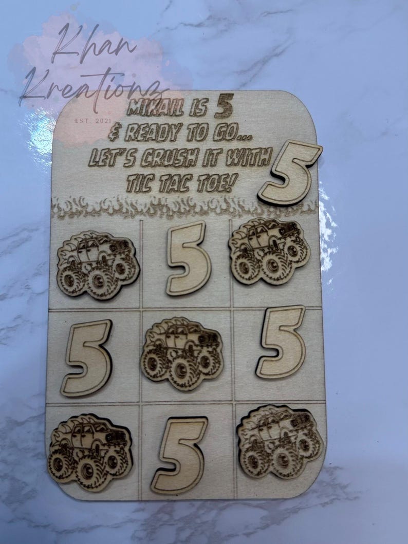 May include: Wooden tic-tac-toe game with laser-cut monster truck and number 5 designs. The game board includes the text "MIKAIL IS 5 & READY TO GO... LET'S CRUSH IT WITH TIC TAC TOE!" and the Khan Kreations logo.