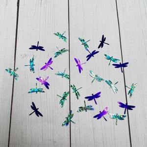 May include: Assortment of iridescent dragonfly cutouts in shades of blue, green, and purple on a white wood background.