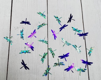 May include: Assortment of iridescent dragonfly cutouts in shades of blue, green, and purple on a white wood background.
