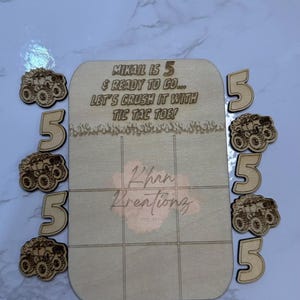 May include: A wooden tic-tac-toe game set with the text "MIKAIL IS 5 & READY TO GO... LET'S CRUSH IT WITH TIC TAC TOE!". Includes wooden number 5s and monster truck cutouts. The game board has a grid design.