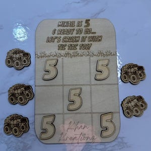 May include: Wooden tic-tac-toe game with the text "MIKAIL IS 5 & READY TO GO... LET'S CRUSH IT WITH TIC TAC TOE!" and the name "Khan Kreationz". Includes wooden number 5s and monster truck game pieces.
