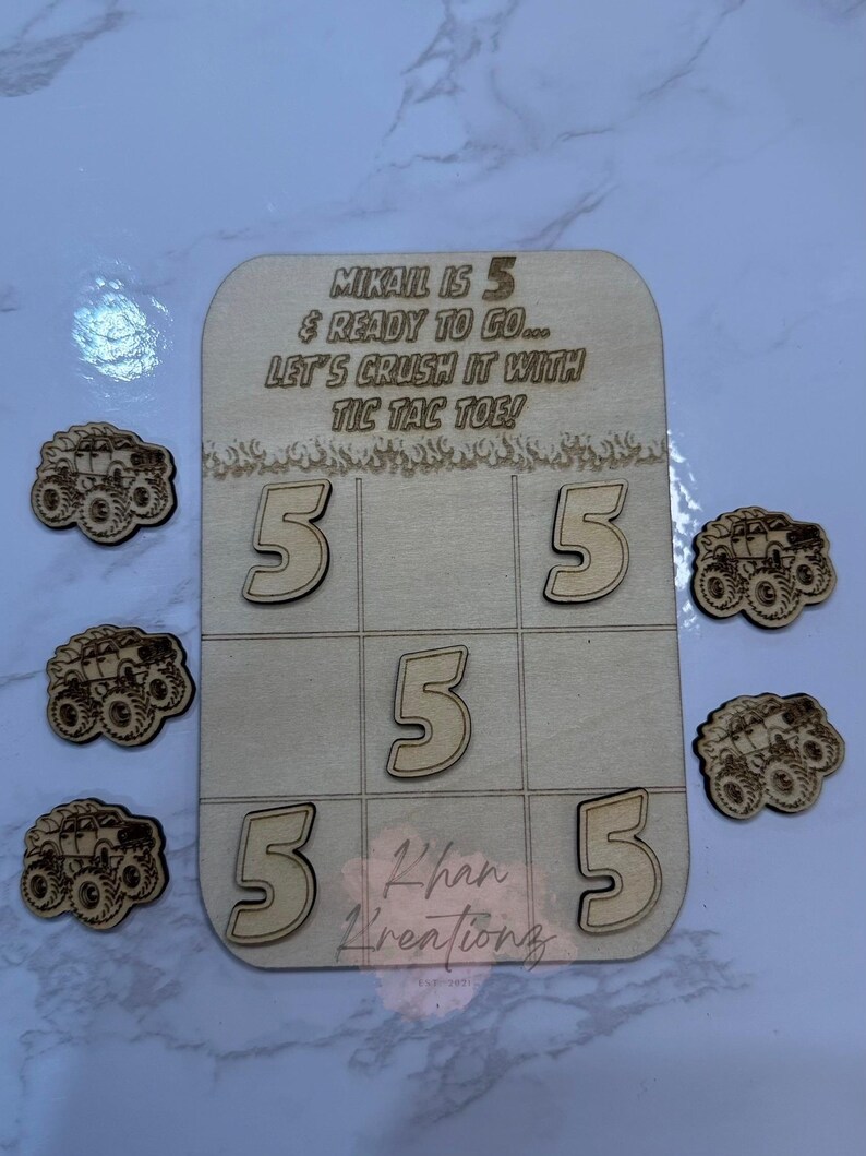 May include: Wooden tic-tac-toe game with the text "MIKAIL IS 5 & READY TO GO... LET'S CRUSH IT WITH TIC TAC TOE!" and the name "Khan Kreationz". Includes wooden number 5s and monster truck game pieces.