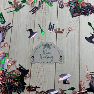May include: A collection of Halloween-themed confetti in black, silver, green, and rose gold colors, including witch hats, brooms, crowns, and wands. The confetti is scattered on a light wood-grain surface, with a sticker that reads "Khan Kreations EST. 2021".