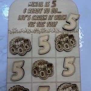 May include: Wooden tic-tac-toe game with the text "My baby turns 5 tomorrow". The game board features the number 5 and monster truck designs. The text "MIKAIL IS 5 & READY TO GO... LET'S CRUSH IT WITH TIC TAC TOE!" is also present.