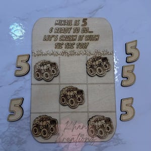 May include: Wooden tic-tac-toe game with monster truck designs. The game board has the text "MIKAIL IS 5 & READY TO GO... LET'S CRUSH IT WITH TIC TAC TOE!" and includes five wooden number 5s. The game pieces are also monster trucks.