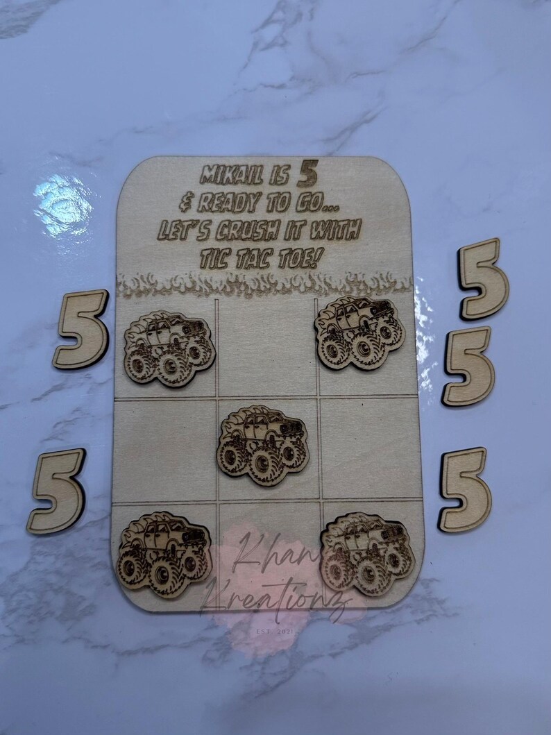 May include: Wooden tic-tac-toe game with monster truck designs. The game board has the text "MIKAIL IS 5 & READY TO GO... LET'S CRUSH IT WITH TIC TAC TOE!" and includes five wooden number 5s. The game pieces are also monster trucks.