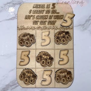 May include: Wooden tic-tac-toe game with a monster truck theme. The game board has the text "MIKAIL IS 5 & READY TO GO... LET'S CRUSH IT WITH TIC TAC TOE!" and includes the number 5 and monster truck graphics.