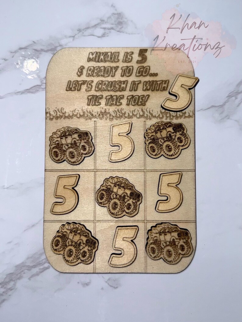 May include: Wooden tic-tac-toe game with a monster truck theme. The game board has the text "MIKAIL IS 5 & READY TO GO... LET'S CRUSH IT WITH TIC TAC TOE!" and includes the number 5 and monster truck graphics.