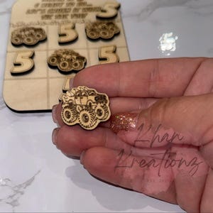 May include: Wooden tic-tac-toe game with laser-cut monster truck pieces and the number 5. The game board has the text "...LET'S CRUISE IT..." and "...THE TIME YOU..." The game pieces are held in a hand.