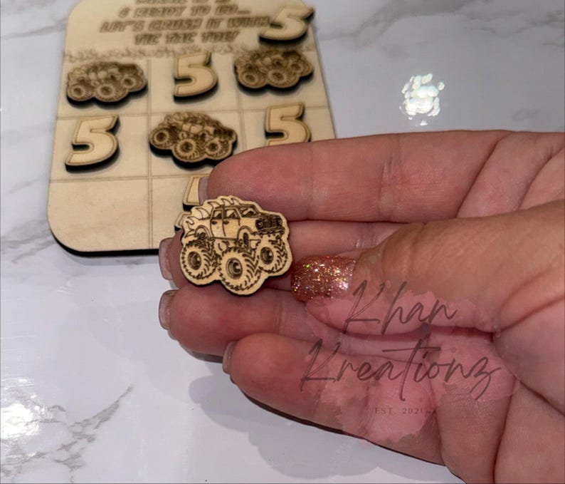 May include: Wooden tic-tac-toe game with laser-cut monster truck pieces and the number 5. The game board has the text "...LET'S CRUISE IT..." and "...THE TIME YOU..." The game pieces are held in a hand.