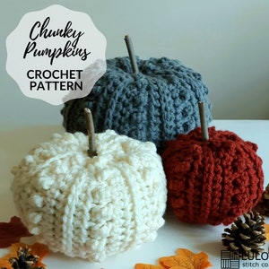 May include: Three crocheted pumpkins in white, gray, and red. The pumpkins are made with a chunky yarn and have a textured surface. The pumpkins are sitting on a white surface with fall leaves and a pine cone.