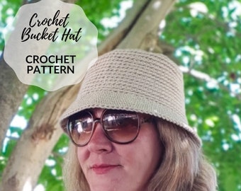 May include: A close-up of a person wearing a beige crochet bucket hat. The hat is crocheted with a textured stitch. The person is wearing sunglasses and has blonde hair. The background is a blurry image of green leaves.