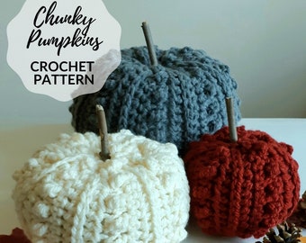 Puede incluir: Tres calabazas de crochet en blanco, gris y rojo. Las calabazas están hechas con un hilo grueso y tienen una superficie texturizada. Las calabazas están sobre una superficie blanca con hojas de otoño y una piña.