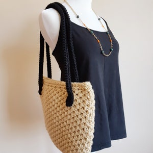 May include: A beige crocheted tote bag with black crocheted straps. The bag is hanging on a white dress form.