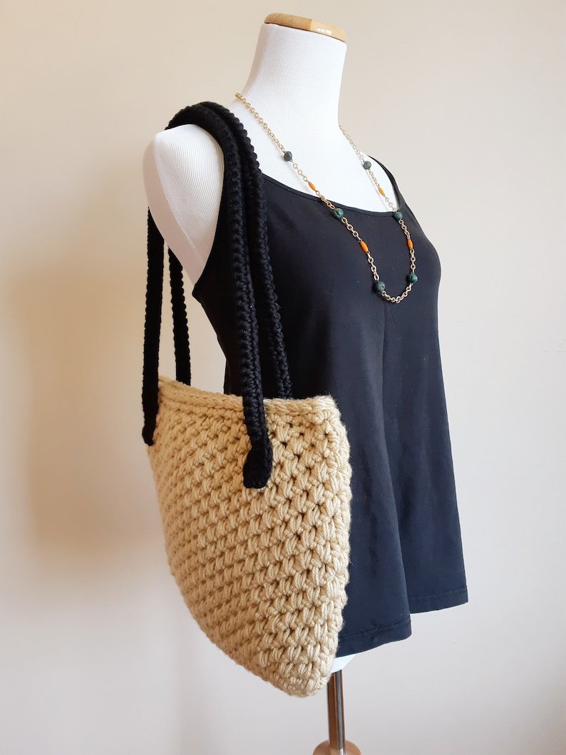 May include: A beige crocheted tote bag with black crocheted straps. The bag is hanging on a white dress form.