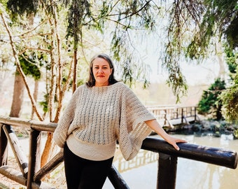 May include: A woman wearing a beige crochet sweater with a lacy pattern, black pants, and black shoes stands on a wooden bridge over a pond. The bridge is made of wooden planks and has a wooden railing. The pond is surrounded by trees and bushes.