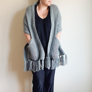 May include: A gray crocheted shawl with fringe and pockets. The shawl is draped over a person's shoulders.