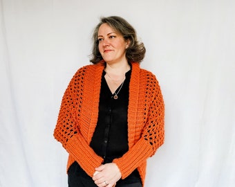 May include: A woman wearing a burnt orange crochet cardigan with a black top and black skirt.