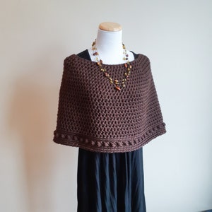 Può includere: Un poncho di uncinetto marrone con un bordo decorativo. Il poncho è esposto su un manichino che indossa un abito nero e una collana con perle ambrate.