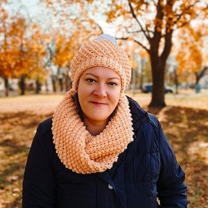 May include: A woman wearing a peach-coloured crocheted hat and scarf. The hat has a white pom-pom on top. She is wearing a dark blue jacket.