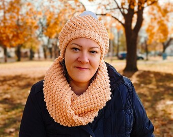 May include: A woman wearing a peach-colored crocheted hat and scarf. The hat has a white pom-pom on top. She is wearing a dark blue jacket.