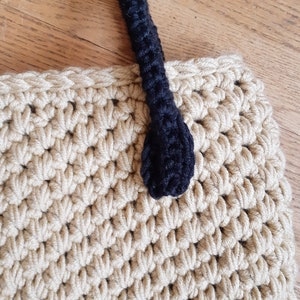 May include: A beige crocheted tote bag with a black crocheted handle. The handle has a rounded end.