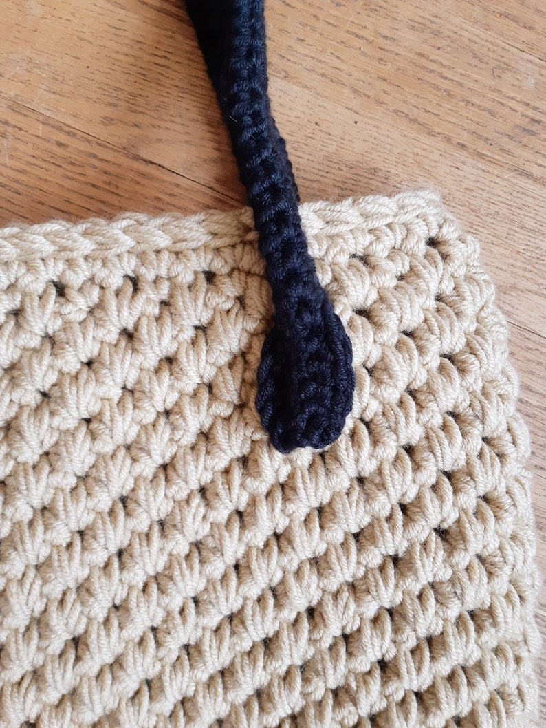 May include: A beige crocheted tote bag with a black crocheted handle. The handle has a rounded end.