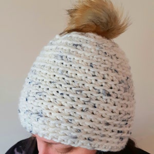 May include: White knitted beanie with a blue fleck pattern and a brown faux fur pom pom on top.