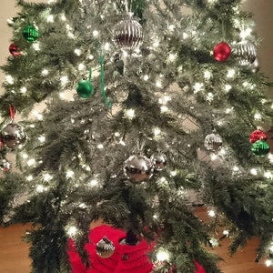 Puede incluir: Una falda de &aacute;rbol de Navidad de punto rojo con botones blancos. La falda se coloca debajo de un &aacute;rbol de Navidad decorado con adornos verdes y plateados y luces blancas.
