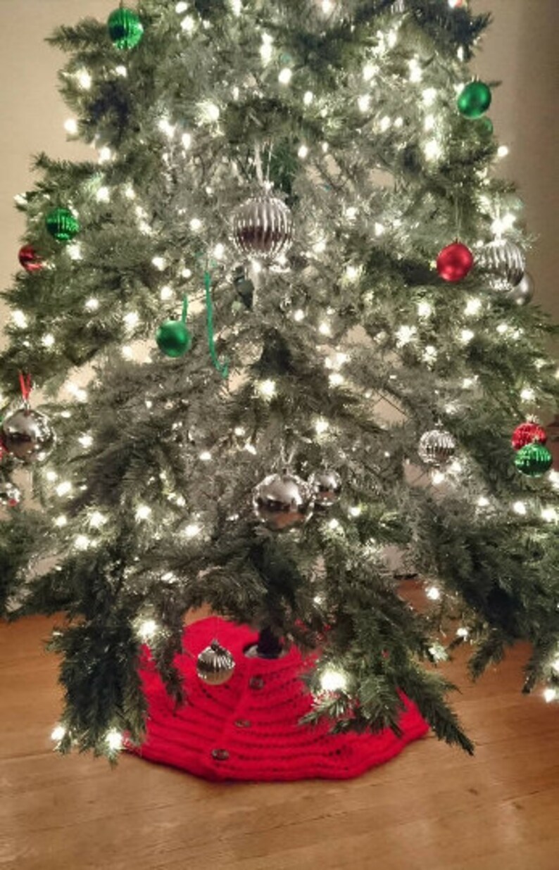 Puede incluir: Una falda de &aacute;rbol de Navidad de punto rojo con botones blancos. La falda se coloca debajo de un &aacute;rbol de Navidad decorado con adornos verdes y plateados y luces blancas.