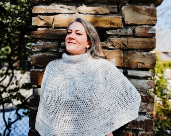 May include: A woman wearing a white crocheted poncho with a textured pattern. She is standing in front of a stone wall.