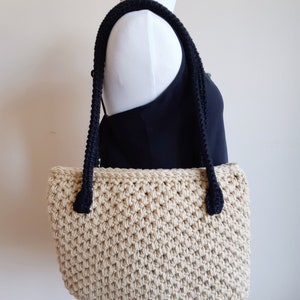 May include: A beige crocheted tote bag with black rope handles. The bag is on a white mannequin.