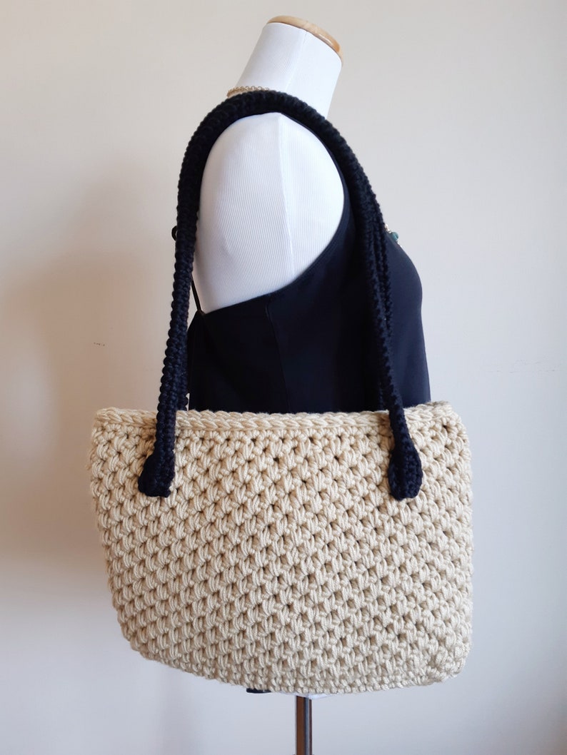 May include: A beige crocheted tote bag with black rope handles. The bag is on a white mannequin.