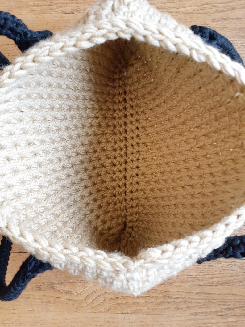 May include: A beige crocheted tote bag with black rope handles. The bag is open and the inside is visible.