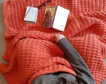 May include: A coral colored crocheted blanket is draped over a bed with a book and a notebook on top. The blanket is made with a textured stitch.