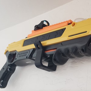 May include: A yellow and black toy gun with a red dot sight and a laser sight. The gun is mounted on a wall with a black bracket.