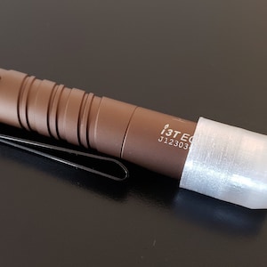 May include: A brown flashlight with a black clip and a white protective sleeve. The flashlight has the text "i3T EC J12303" printed on the side.